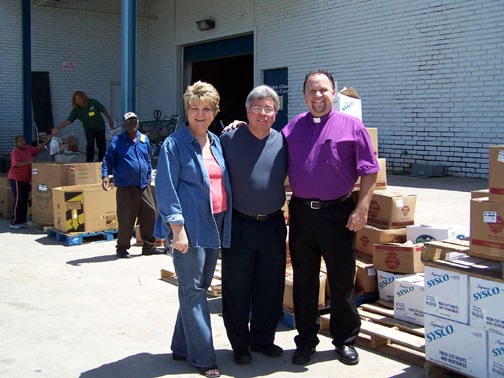 Three people at a dock beside cases of food, one person in a clerical shirt.